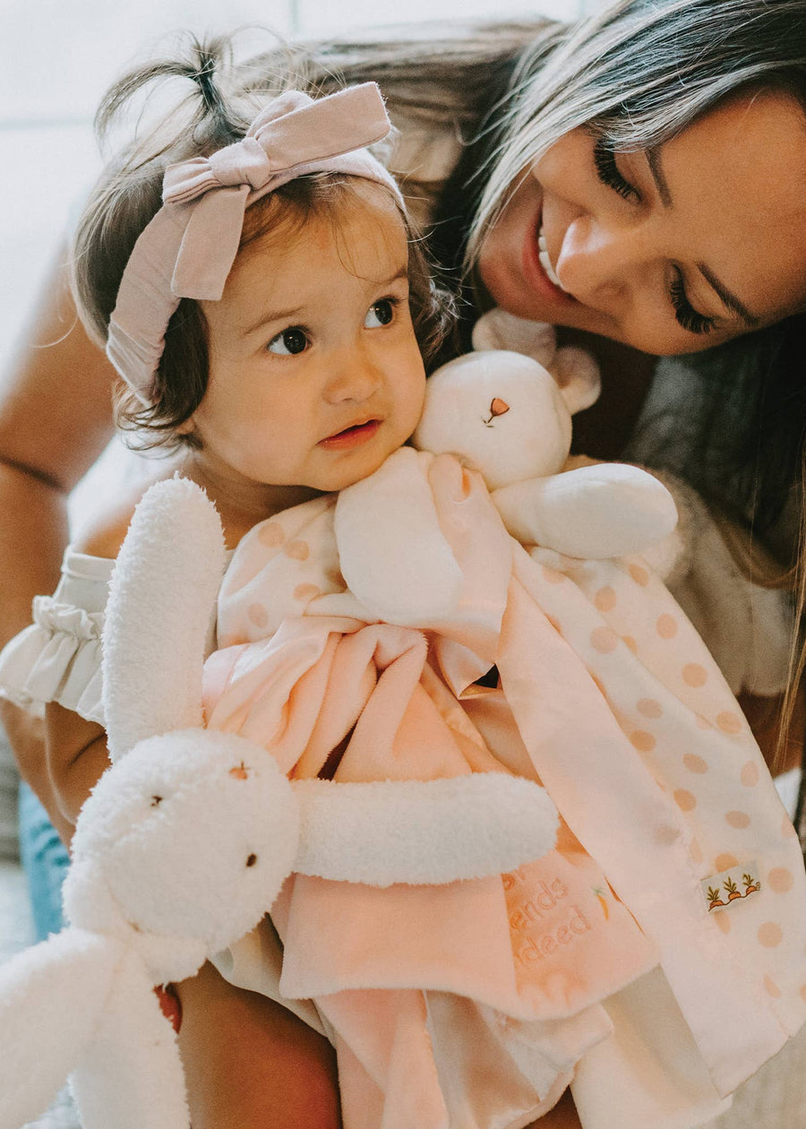 Woman holding a baby dressed in a pink outfit with a bow, surrounded by soft toys.