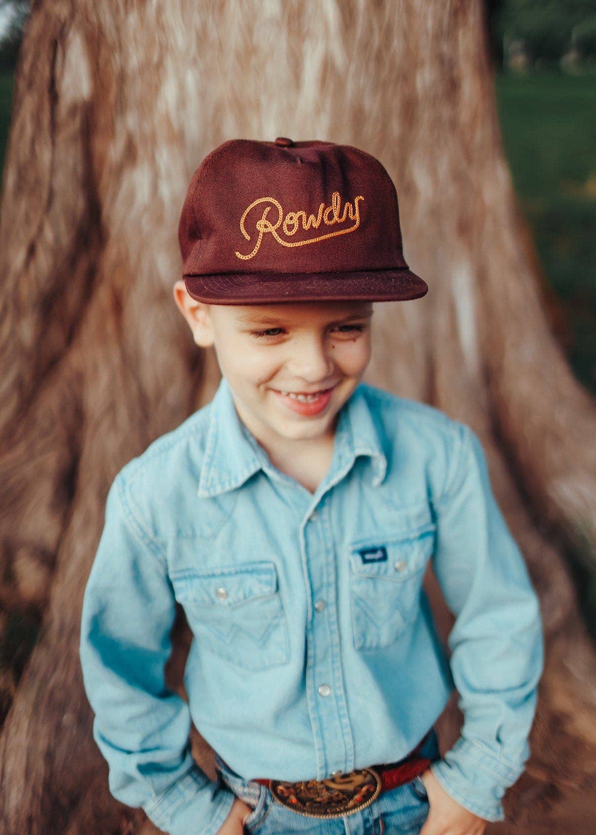 Child wearing a maroon cap with 'Rowdy' text, light blue shirt, and jeans standing in front of a tree.