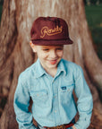 Child wearing a maroon cap with 'Rowdy' text, light blue shirt, and jeans standing in front of a tree.