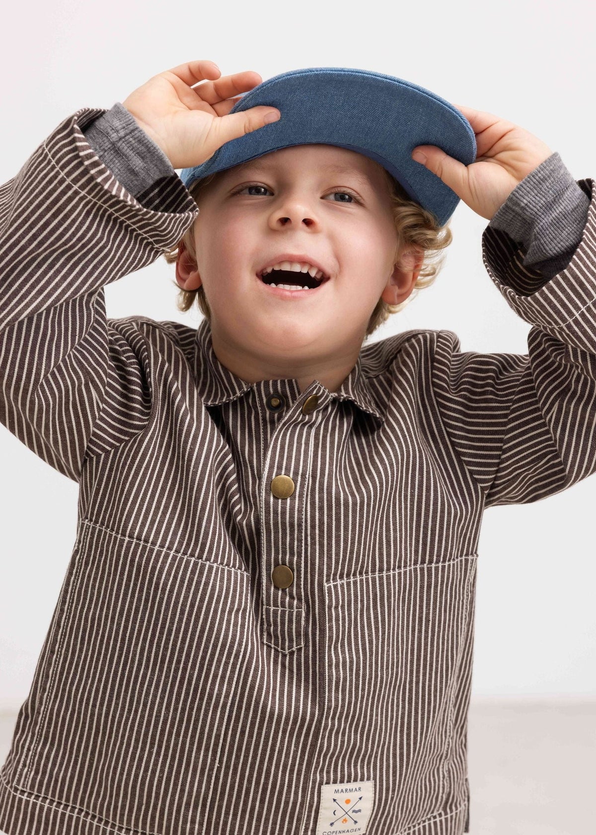 A young boy wearing a brown and beige vertically striped long-sleeve shirt with a blue cap, smiling and adjusting the cap against a plain light background.