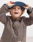 A young boy wearing a brown and beige vertically striped long-sleeve shirt with a blue cap, smiling and adjusting the cap against a plain light background.