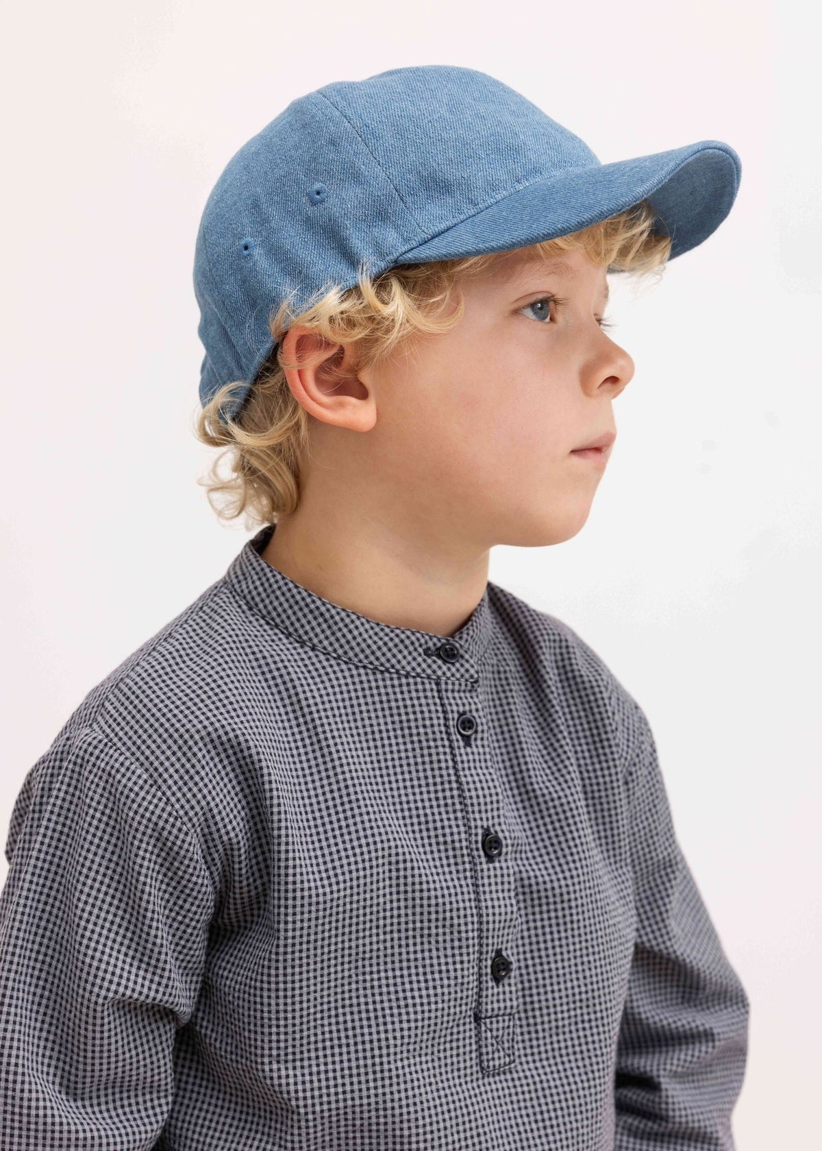 A young child in profile wearing a light blue denim baseball cap and a gray-and-white checkered long-sleeve shirt, photographed against a clean white background.