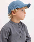 A young child in profile wearing a light blue denim baseball cap and a gray-and-white checkered long-sleeve shirt, photographed against a clean white background.