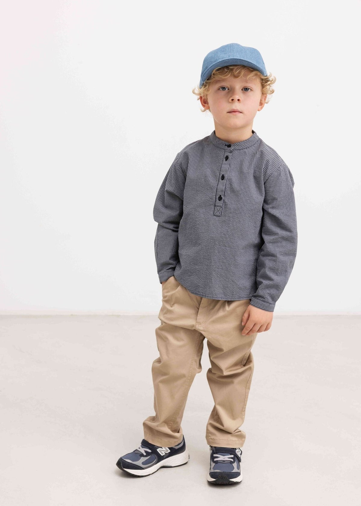 A toddler boy wearing a gray long-sleeve henley shirt, beige pants, navy sneakers, and a blue cap, standing against a plain white studio background.