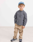 A toddler boy wearing a gray long-sleeve henley shirt, beige pants, navy sneakers, and a blue cap, standing against a plain white studio background.