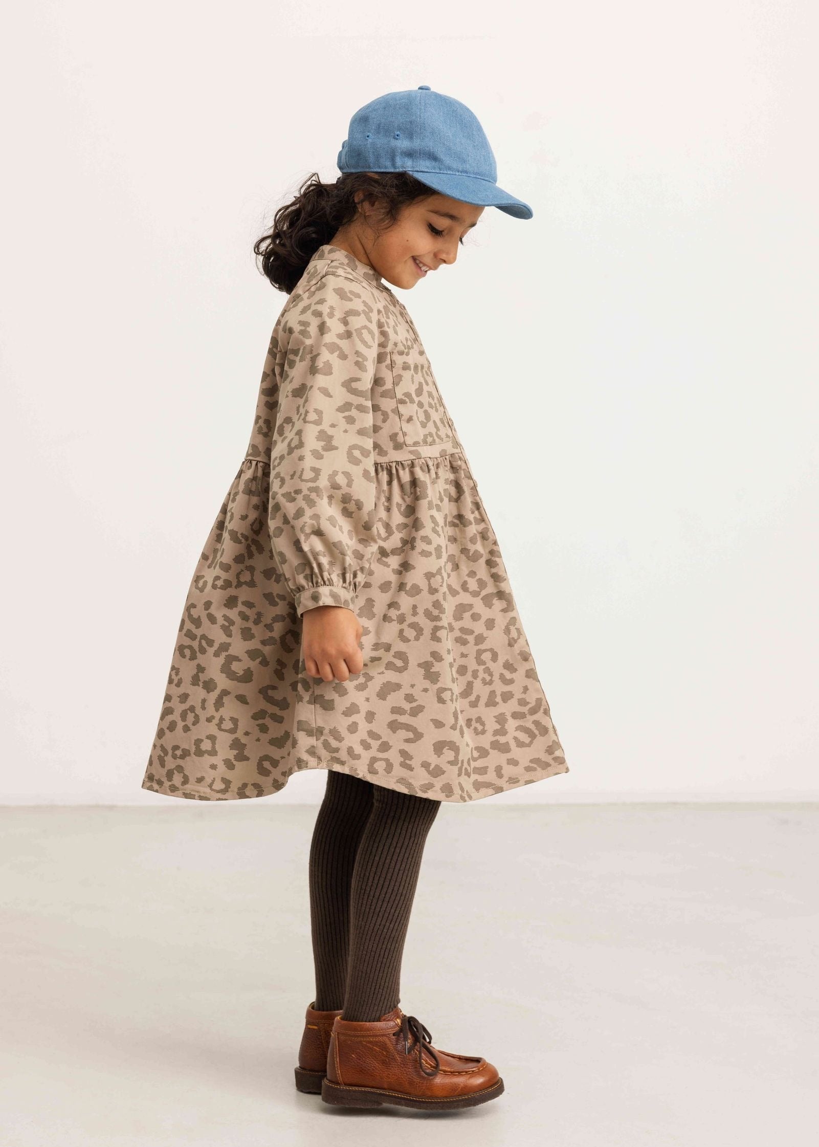 A young child wearing a beige and brown leopard-print dress, a light blue cap, dark brown tights, and brown leather shoes, standing in profile against a plain white studio background.