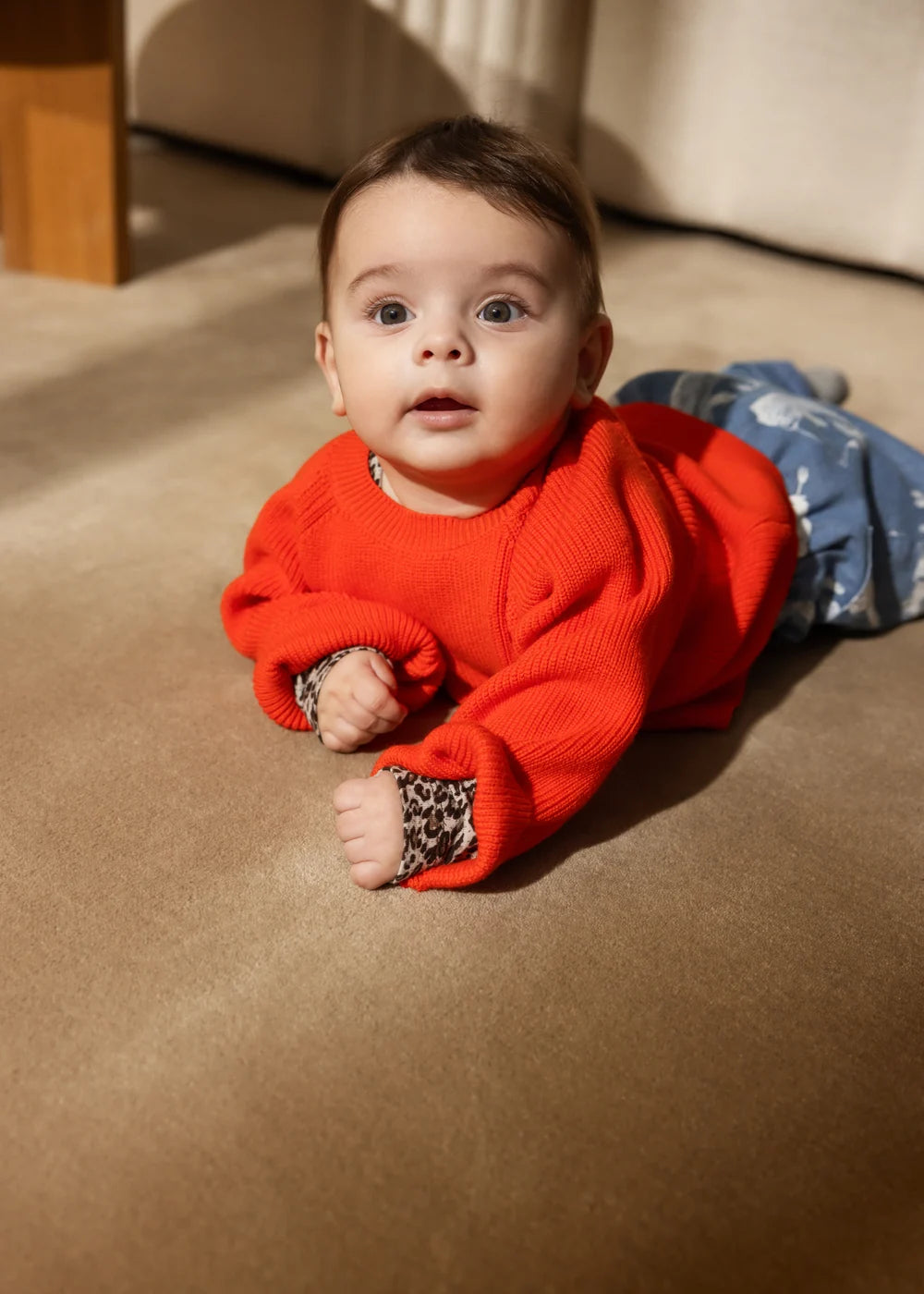 A baby lying on a beige carpet wearing a bright orange-red knit sweater with leopard-print cuffs and blue patterned pants.