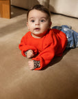 A baby lying on a beige carpet wearing a bright orange-red knit sweater with leopard-print cuffs and blue patterned pants.
