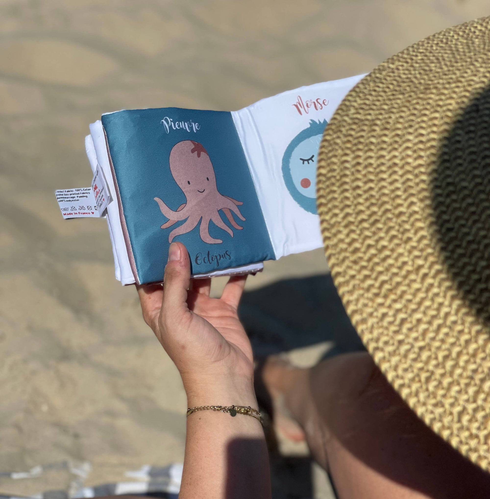 A person at the beach holds an open small book or journal. The cover is teal with a pink octopus illustration and the title 'Pipie Clippings.