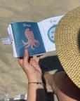 A person at the beach holds an open small book or journal. The cover is teal with a pink octopus illustration and the title 'Pipie Clippings.