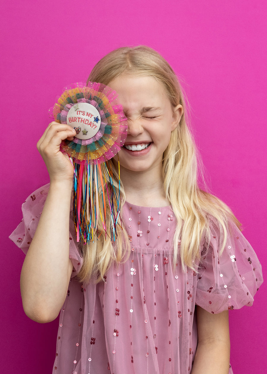 Young girl in a pink dress holding a colorful birthday badge against a pink background