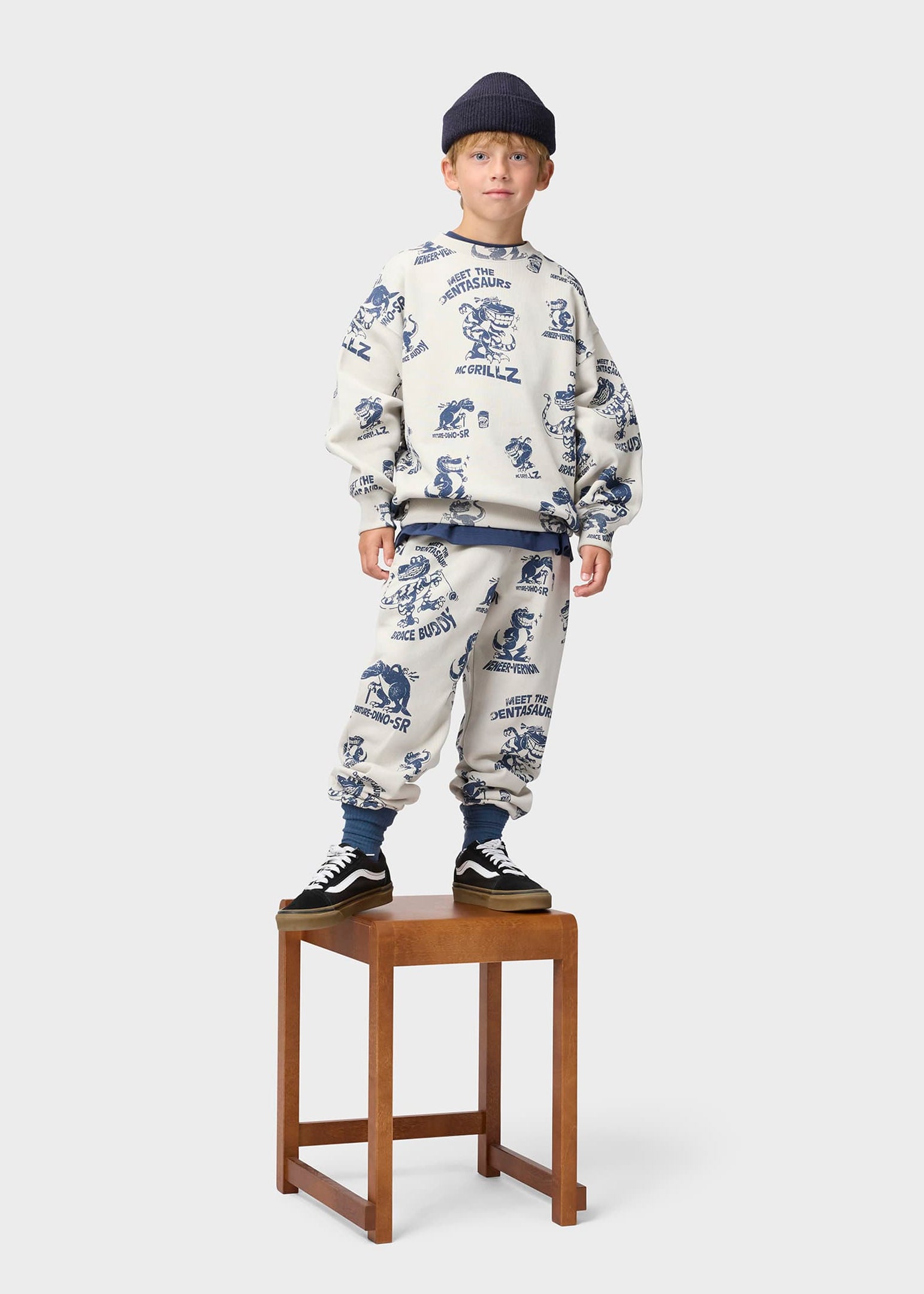Child wearing a patterned outfit standing on a wooden stool against a white background