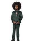 A child model wearing a dark green two-piece suit with a patterned shirt and tie, standing against a light gray studio background.