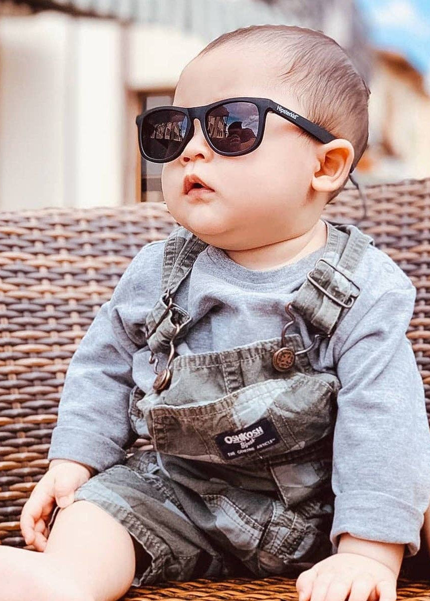 Baby wearing sunglasses and overalls sitting on a wicker chair.