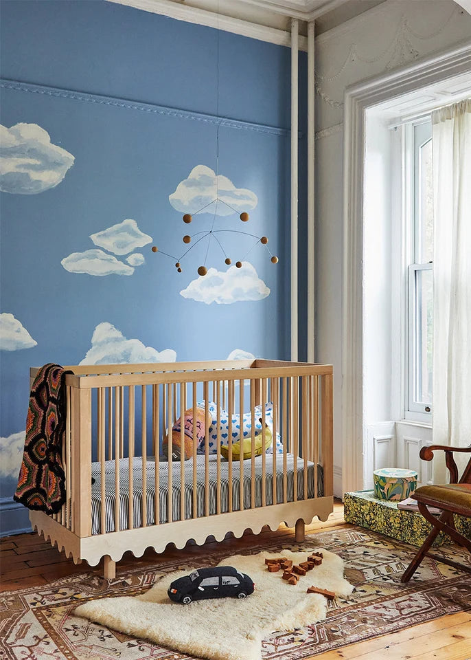 Nursery room with blue wall featuring cloud mural, wooden crib, and decorative chair.