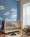 Nursery room with blue wall featuring cloud mural, wooden crib, and decorative chair.
