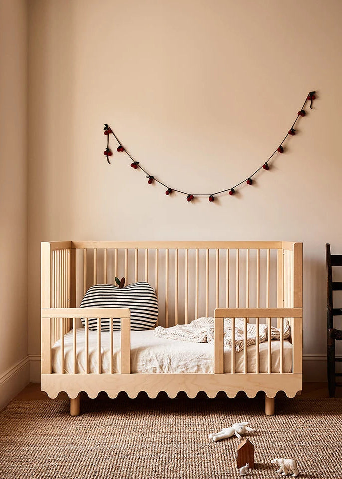 A light natural wood toddler bed with safety rails, styled in a warm beige nursery with a striped pillow, woven rug, and a decorative string of lights on the wall.