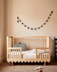 A light natural wood toddler bed with safety rails, styled in a warm beige nursery with a striped pillow, woven rug, and a decorative string of lights on the wall.