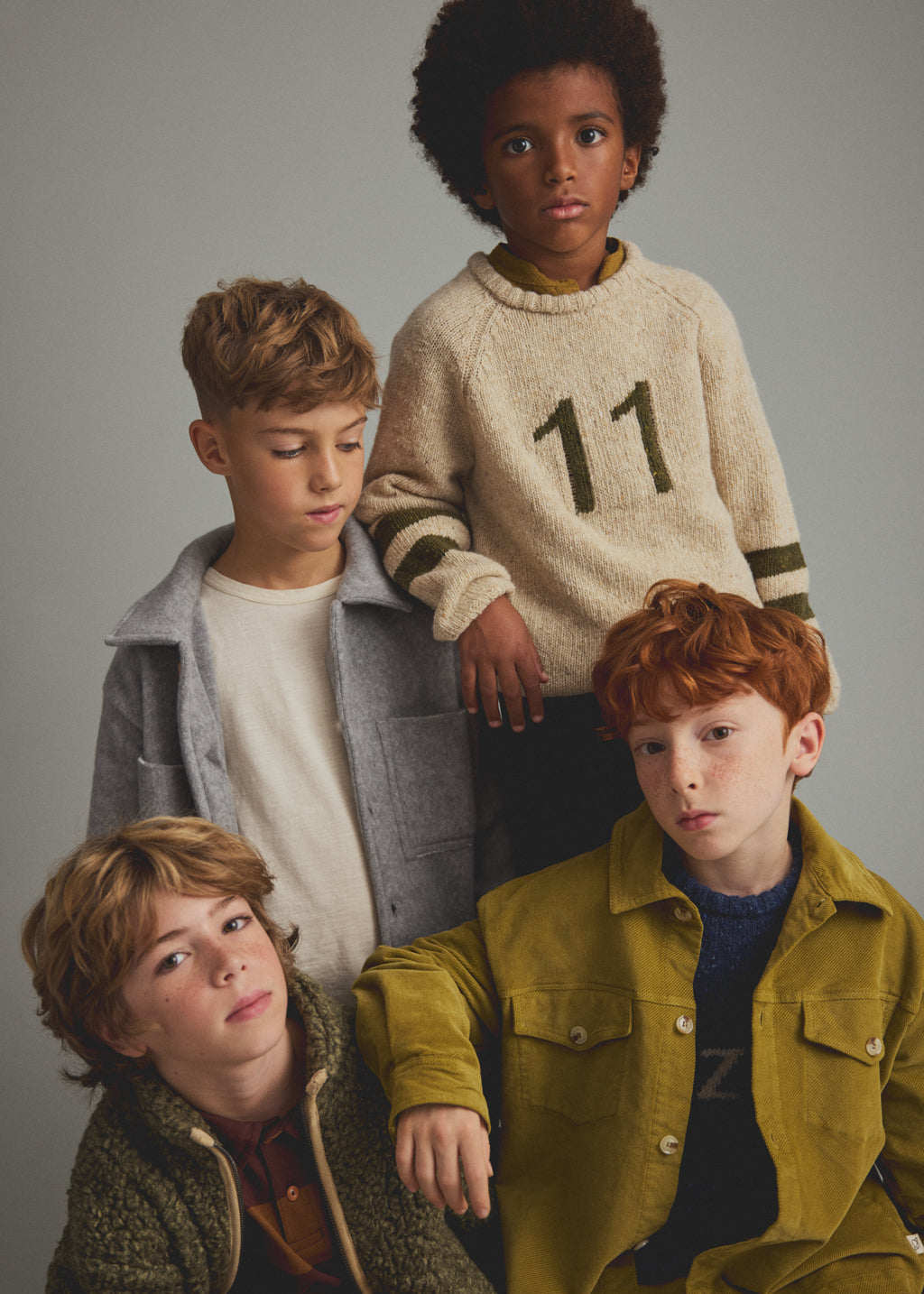Four boys modeling knit and casual outerwear in a studio setting against a solid gray background. The group includes a boy in a cream sweater with the number 11, a boy in a gray jacket over a white shirt, a boy in a green corduroy jacket, and a boy in a mustard green corduroy jacket over a dark shirt.