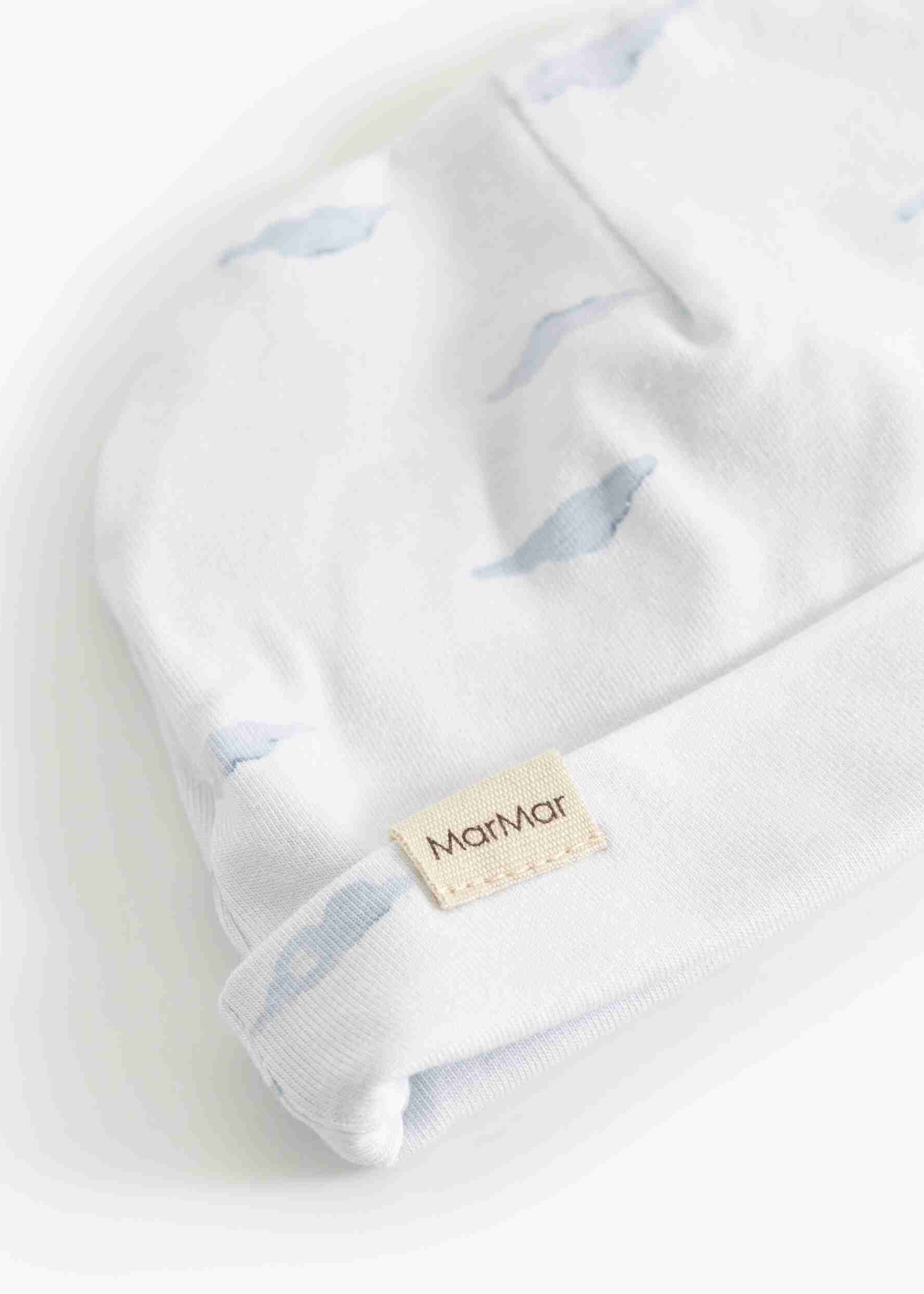 A close-up of a white knit baby hat with a soft blue whale print and a beige brand tag reading MartMar.