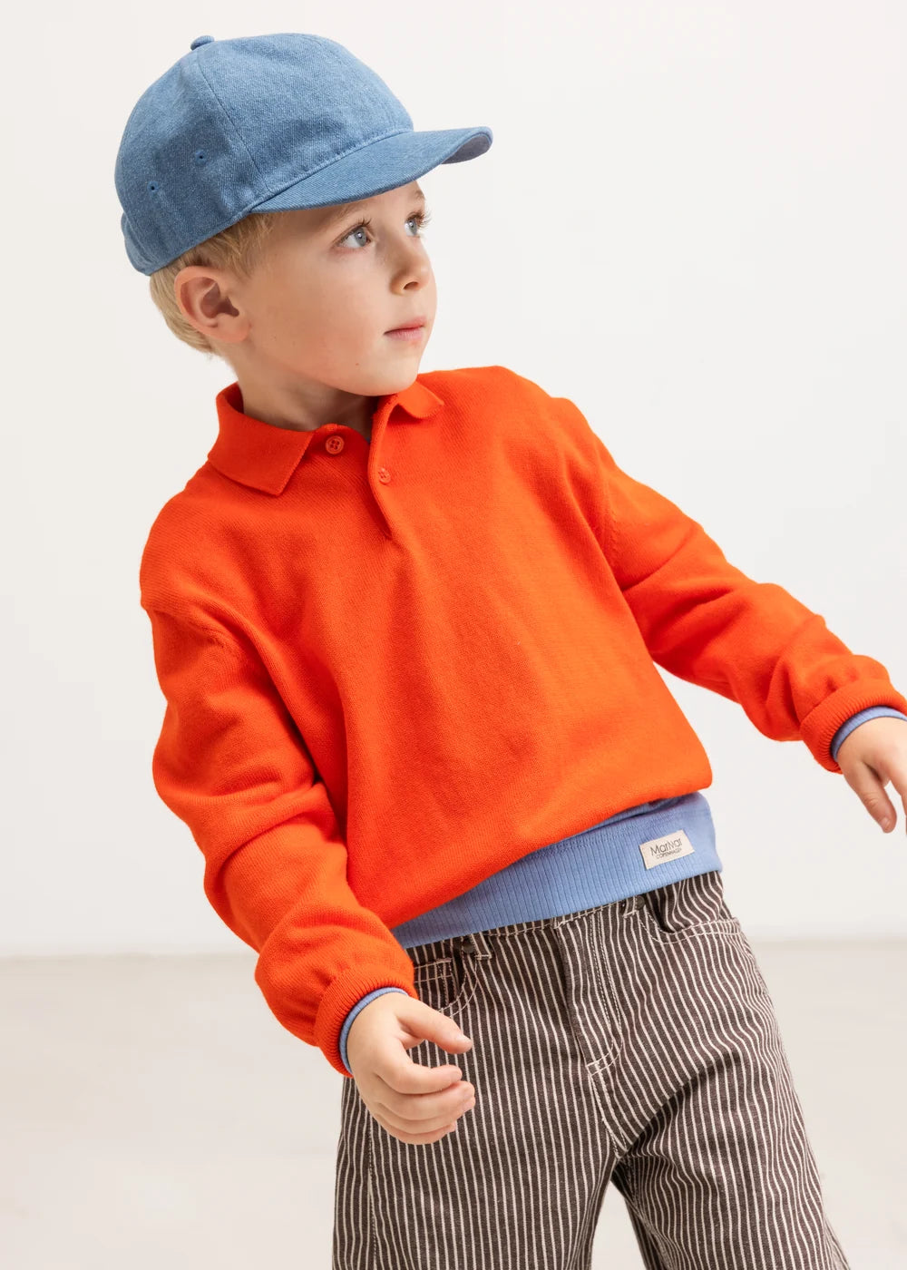 Child wearing an orange sweater, blue cap, and striped pants on a white background