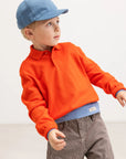 Child wearing an orange sweater, blue cap, and striped pants on a white background
