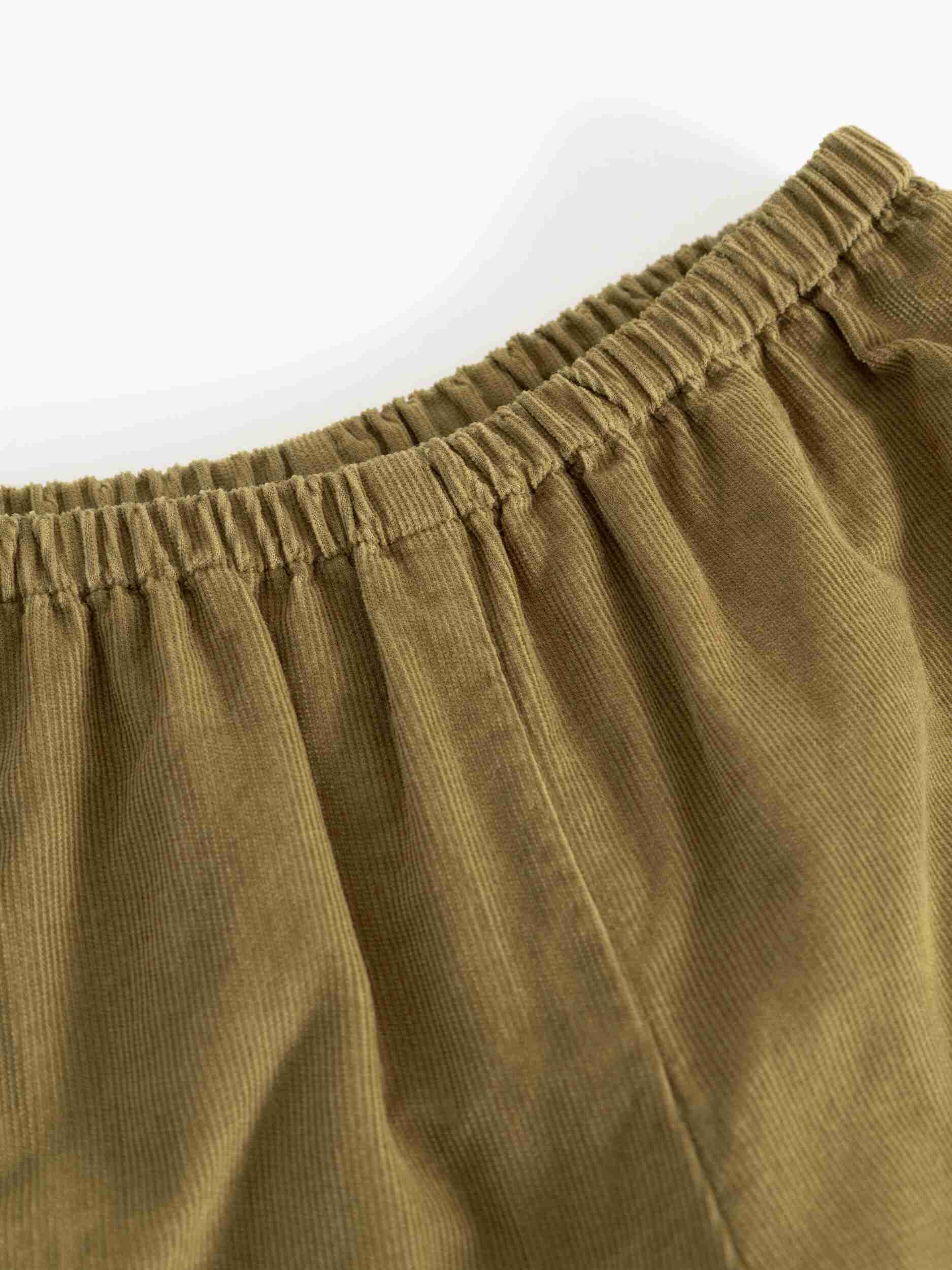 Close-up of a corduroy garment in a muted olive-brown color, showing the elastic waistband and ribbed fabric texture against a plain white background.