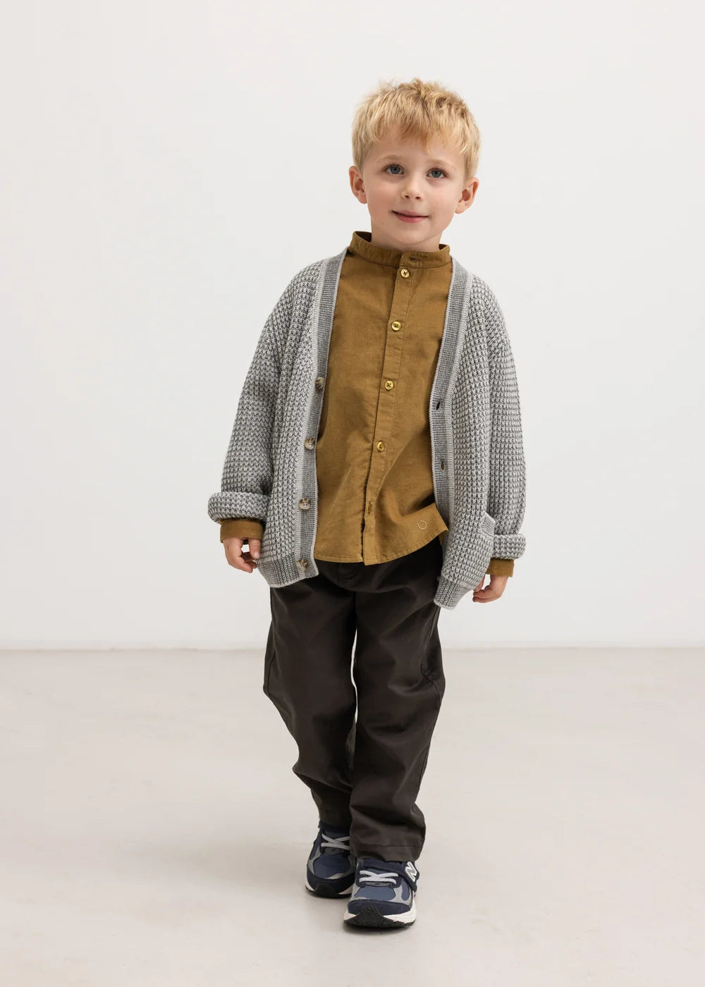 Young boy wearing a brown shirt, gray cardigan, and dark pants on a white background