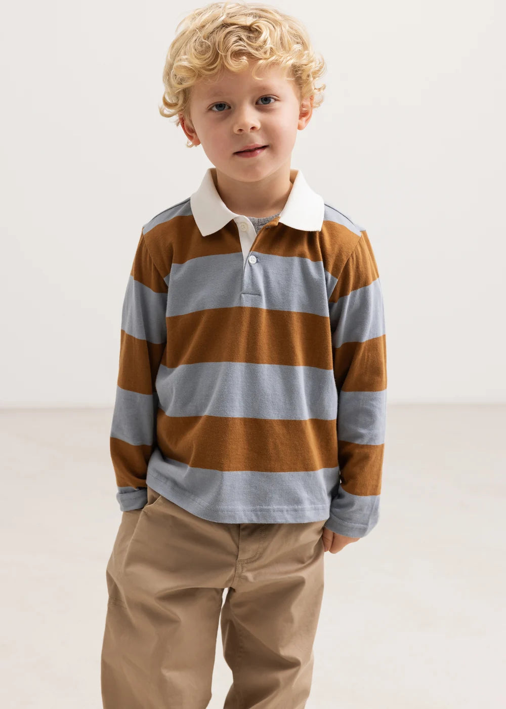 A young boy with curly blonde hair wearing a long-sleeve rugby shirt with brown and light gray horizontal stripes, paired with khaki pants, standing against a plain light background.