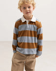 A young boy with curly blonde hair wearing a long-sleeve rugby shirt with brown and light gray horizontal stripes, paired with khaki pants, standing against a plain light background.
