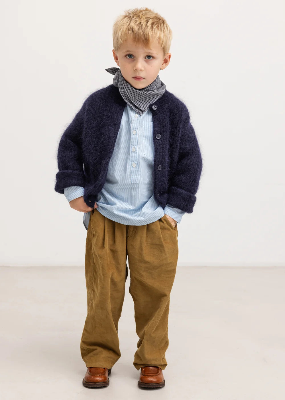 Young boy wearing a navy cardigan, light blue shirt, brown pants, and brown shoes on a white background