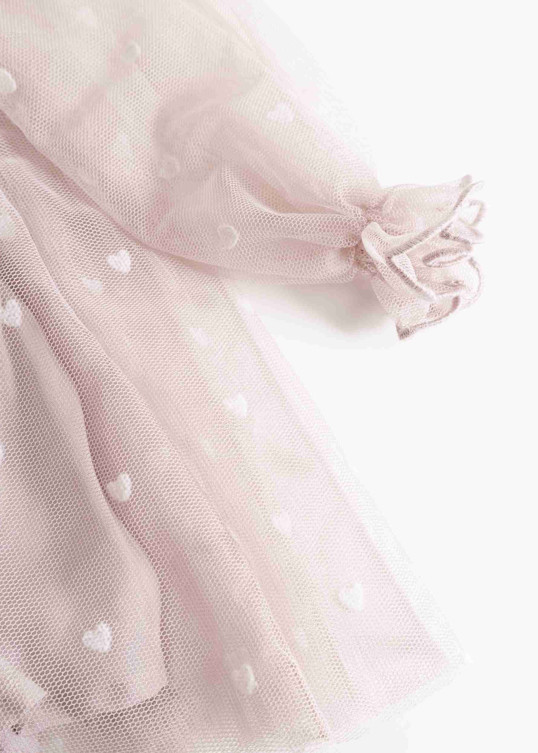 Close-up of a light pink tulle skirt with small white heart motifs and a ruffled tie detail on the sleeve, shown against a plain white background.