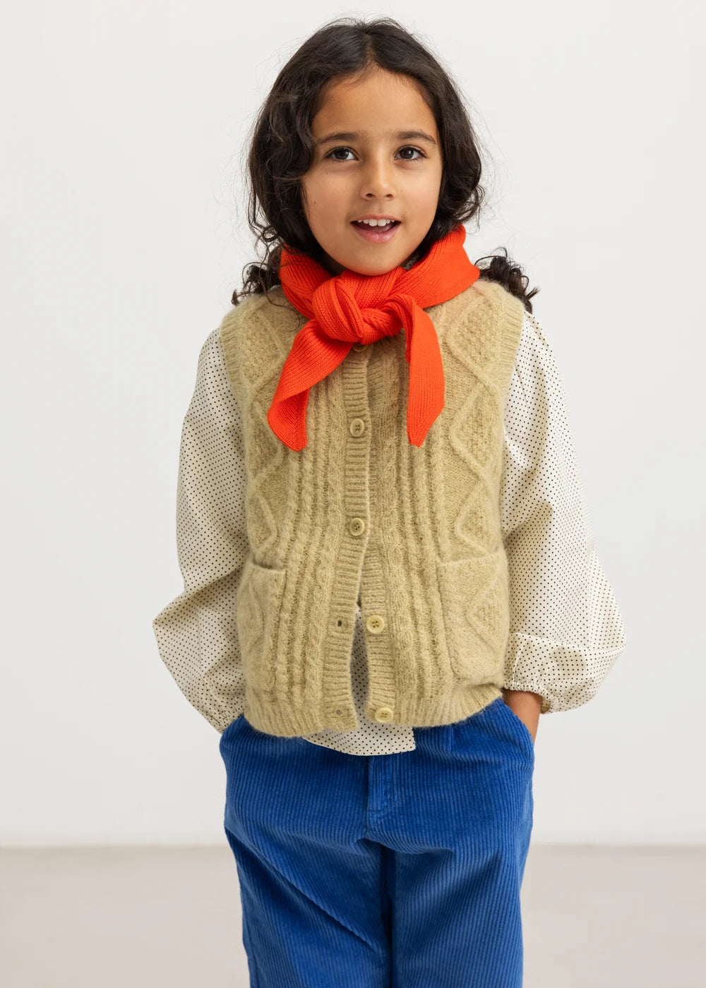 A young girl with dark curly hair stands against a plain white studio background, wearing a cream-colored textured knit cardigan vest over a white long-sleeve shirt, a bright orange knitted scarf tied in a bow, and blue corduroy pants.