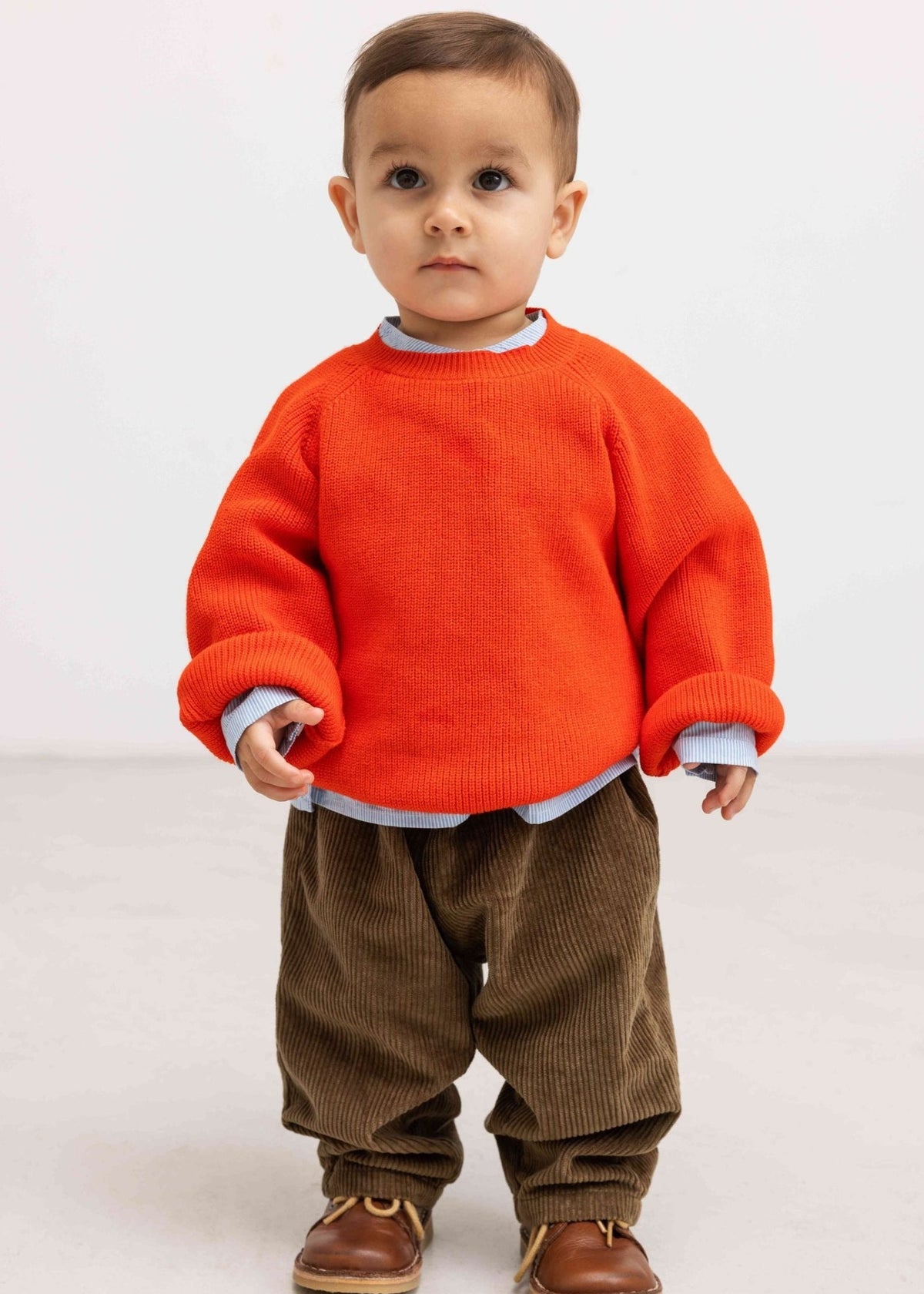 A toddler standing against a plain light gray studio background, wearing a bright orange knit sweater over a light blue collared shirt, brown corduroy pants, and brown shoes.