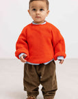 A toddler standing against a plain light gray studio background, wearing a bright orange knit sweater over a light blue collared shirt, brown corduroy pants, and brown shoes.