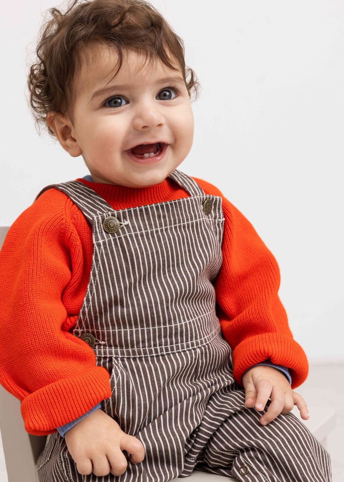 Child wearing a red shirt and brown striped overalls sitting on a white surface.