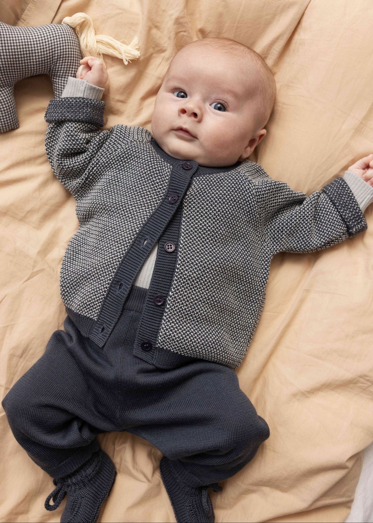 A baby lying on beige bedding, wearing a gray textured knit cardigan over a white undershirt and dark navy pants.