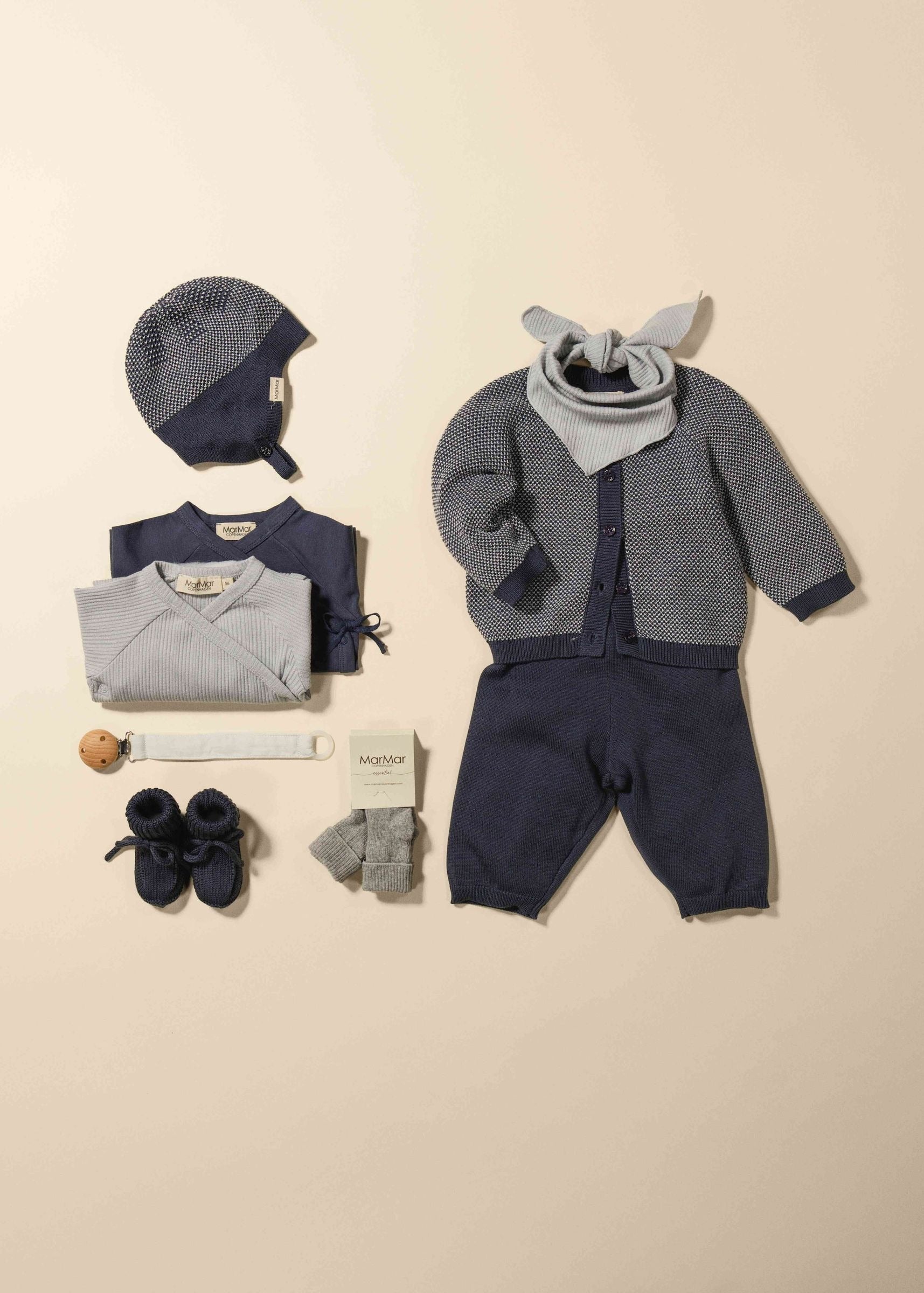 A baby clothing set laid out on a warm beige background, including a navy and gray knit bonnet, folded top, light gray wrap, cardigan with gray bunny ears, navy pants, gray socks, navy booties, and a white bandana.