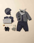 A baby clothing set laid out on a warm beige background, including a navy and gray knit bonnet, folded top, light gray wrap, cardigan with gray bunny ears, navy pants, gray socks, navy booties, and a white bandana.