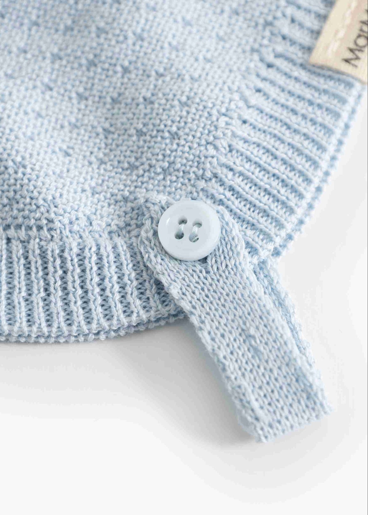 Close-up of a light blue knit baby garment with a ribbed cuff, a matching light blue button tab, and part of a beige fabric tag.