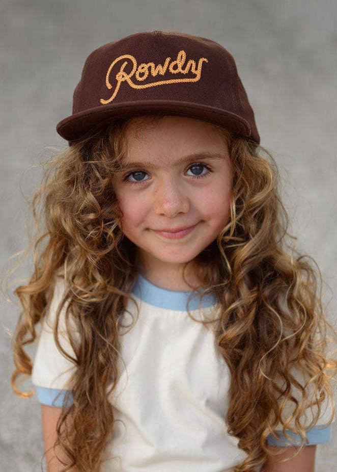 Young girl wearing a brown cap with 'Rowdy' embroidered on it against a gray background