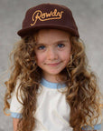 Young girl wearing a brown cap with 'Rowdy' embroidered on it against a gray background