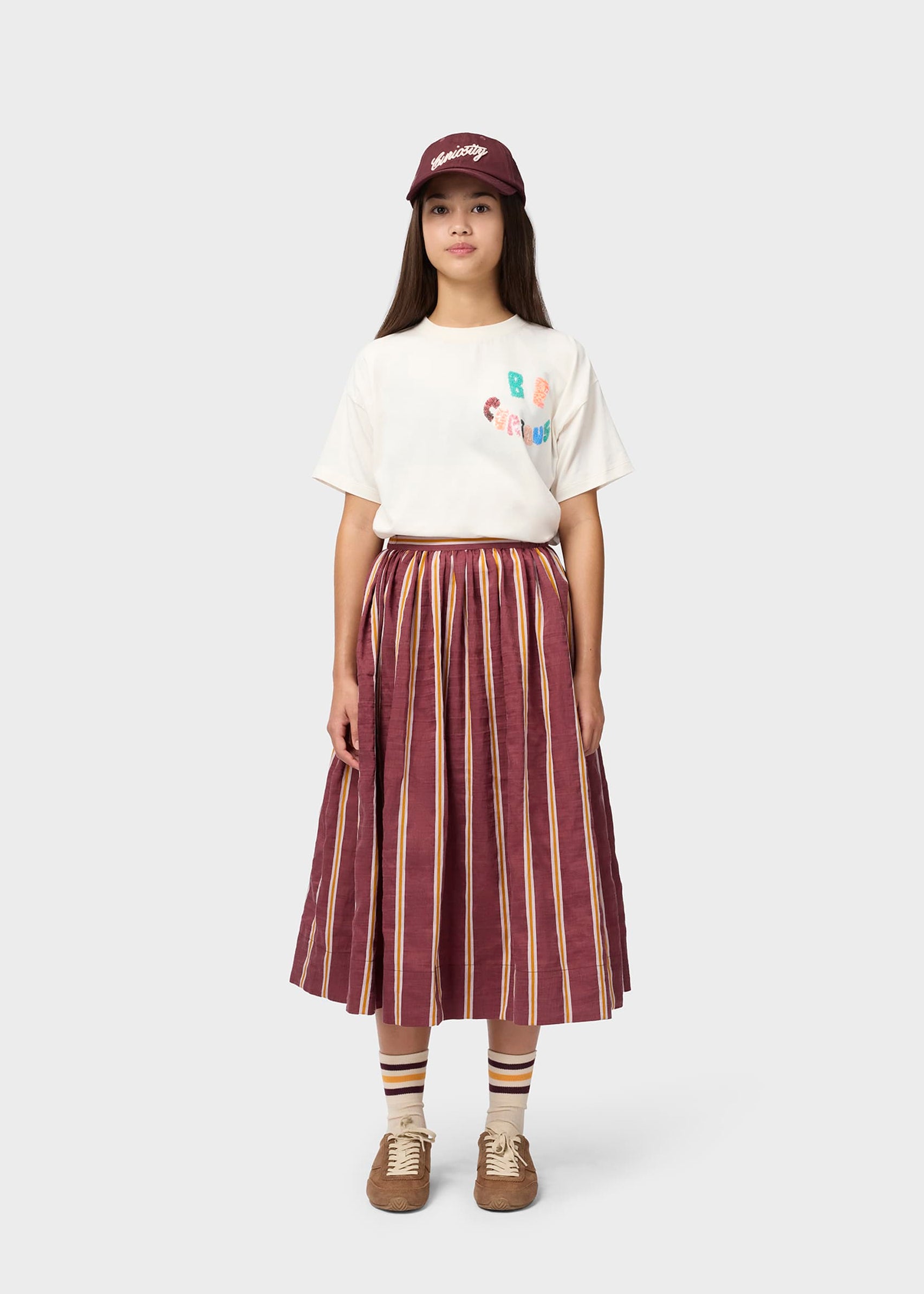 Person wearing a white t-shirt with colorful graphics, a maroon and beige striped skirt, and a maroon cap on a white background.