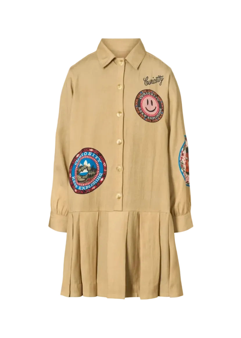 A beige long-sleeve button-up shirt dress with a pleated hem, featuring colorful embroidered patches and 'Curiosity' script on the chest, shown against a plain white background.