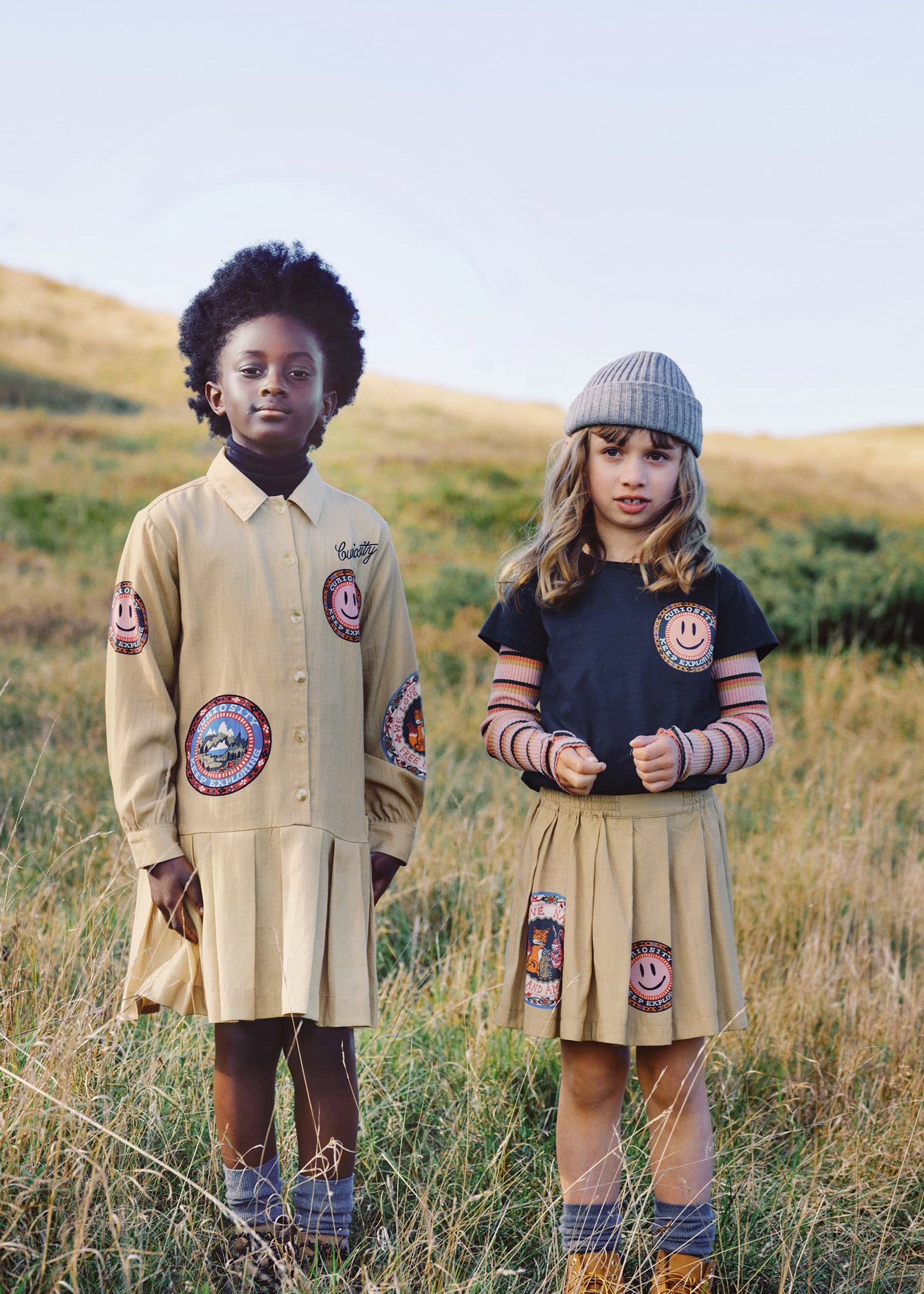 Two children standing in an outdoor grassy field wearing matching beige patchwork dresses and a navy top with striped sleeves, both featuring circular embroidered patches.