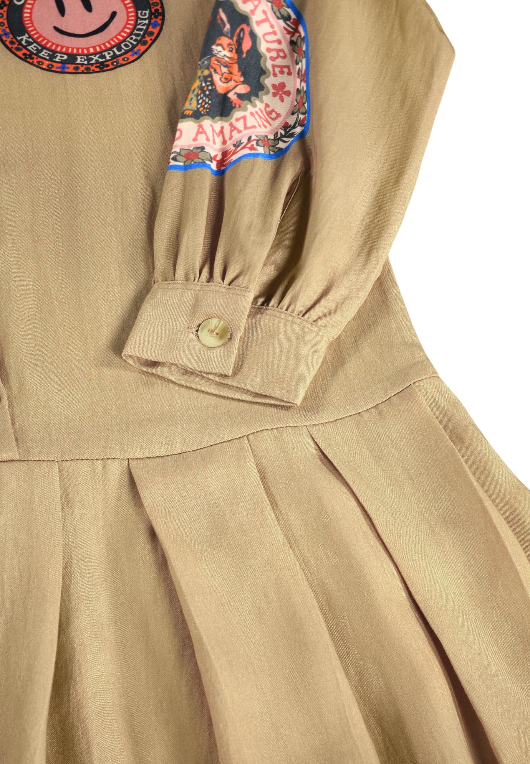 Close-up of a beige pleated dress with a buttoned cuff, featuring colorful embroidered patches on the chest.