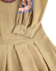 Close-up of a beige pleated dress with a buttoned cuff, featuring colorful embroidered patches on the chest.