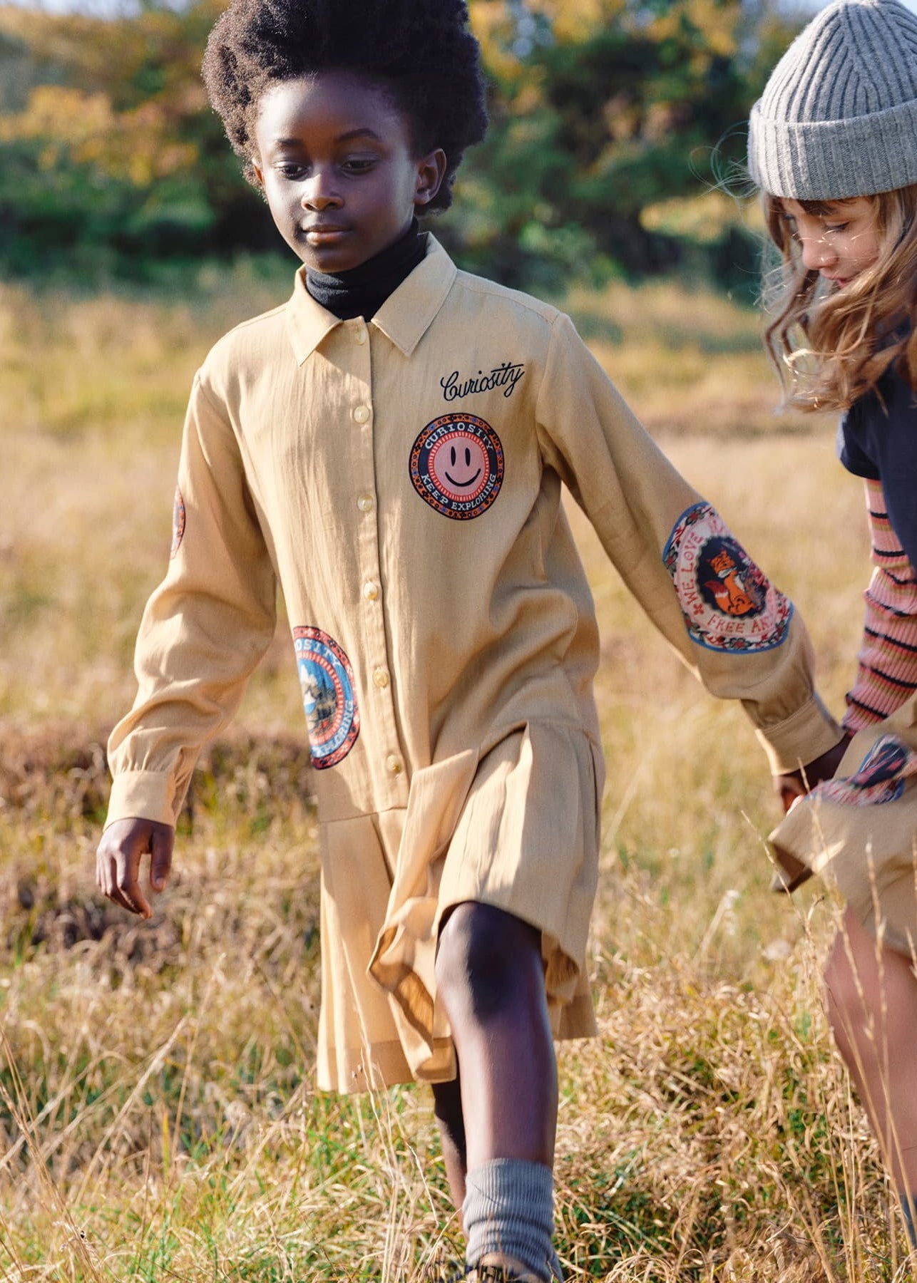 Two children outdoors in a grassy field, wearing matching tan scout-style uniforms with circular embroidered patches. One child is standing and the other is crouching, both looking down at the ground.