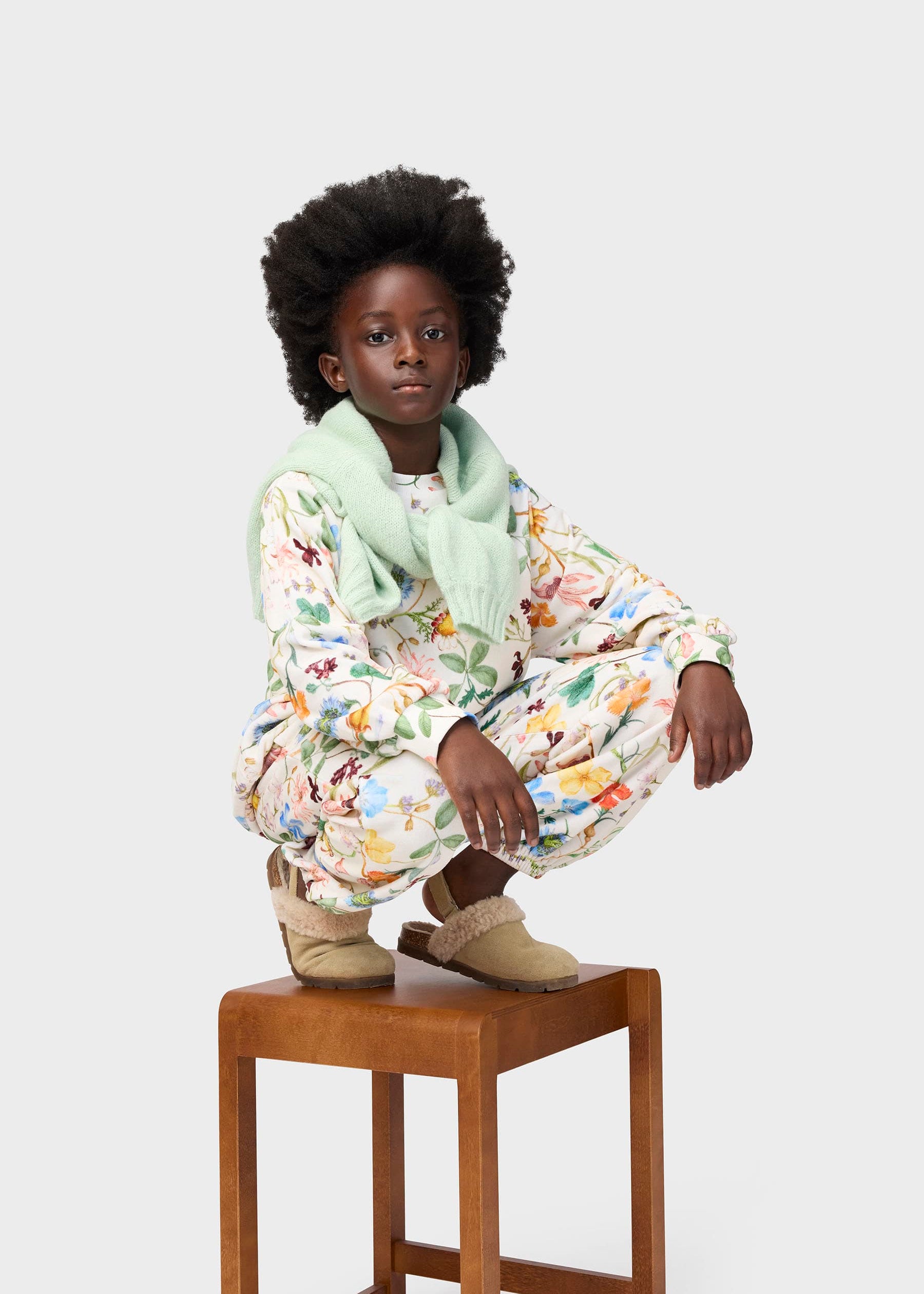 Child wearing a floral outfit with a green scarf, sitting on a wooden stool against a white background