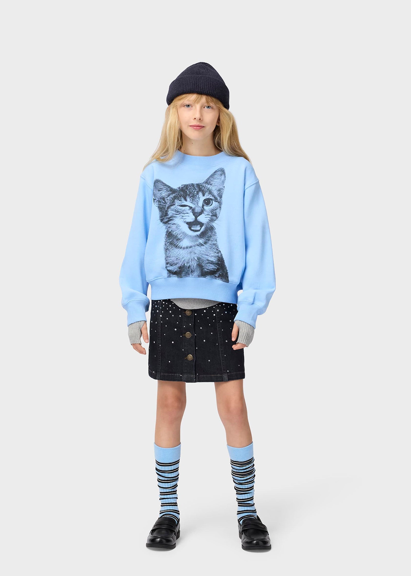 Child wearing a light blue sweatshirt with a cat print, black skirt, and striped socks on a white background